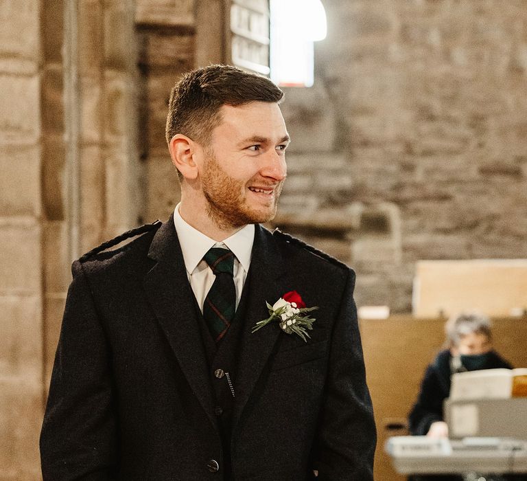 Groom awaits his bride wearing three piece suit complete with kilt and white and red floral buttonhole 