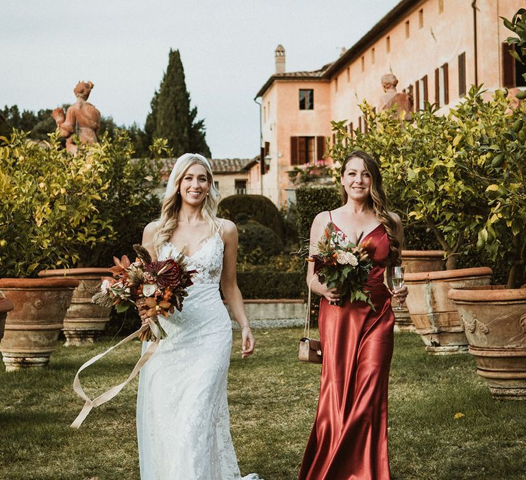 Bride stands with her bridesmaid wearing silk burnt orange bridesmaid dress with thin straps and low cut front 