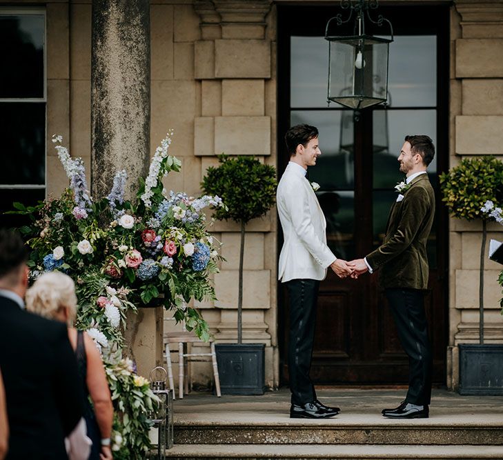 Grooms wearing black tie stand between pastel coloured large floral bouquets at Birdsall House during outdoor ceremony