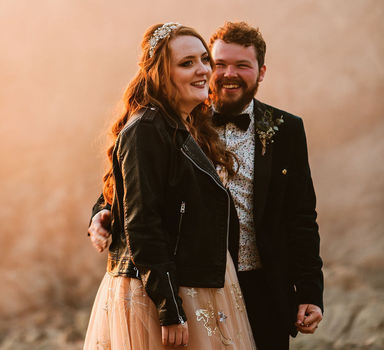 Bride wears personalised leather jacket and pearl embellished wedding crown whilst stood with her groom within orange smoke bomb