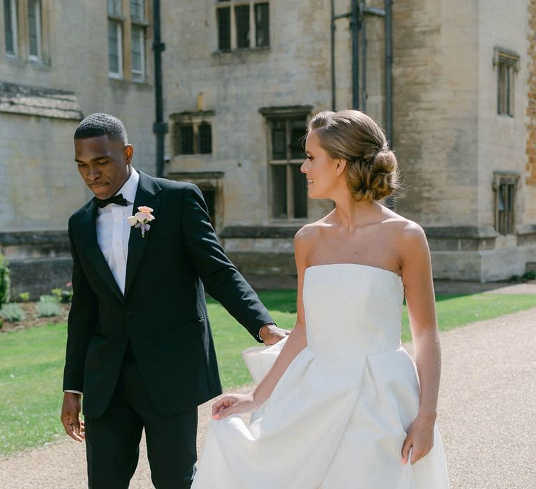 Bride in strapless Jesus Peiro wedding dress walking with the groom in black tie around the grounds of Rushton Hall 