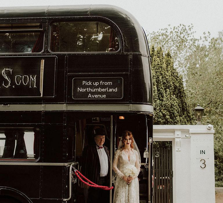 Bride stands on black London Ghost Bus on the morning of her wedding day beside driver in top hat and black tie dress