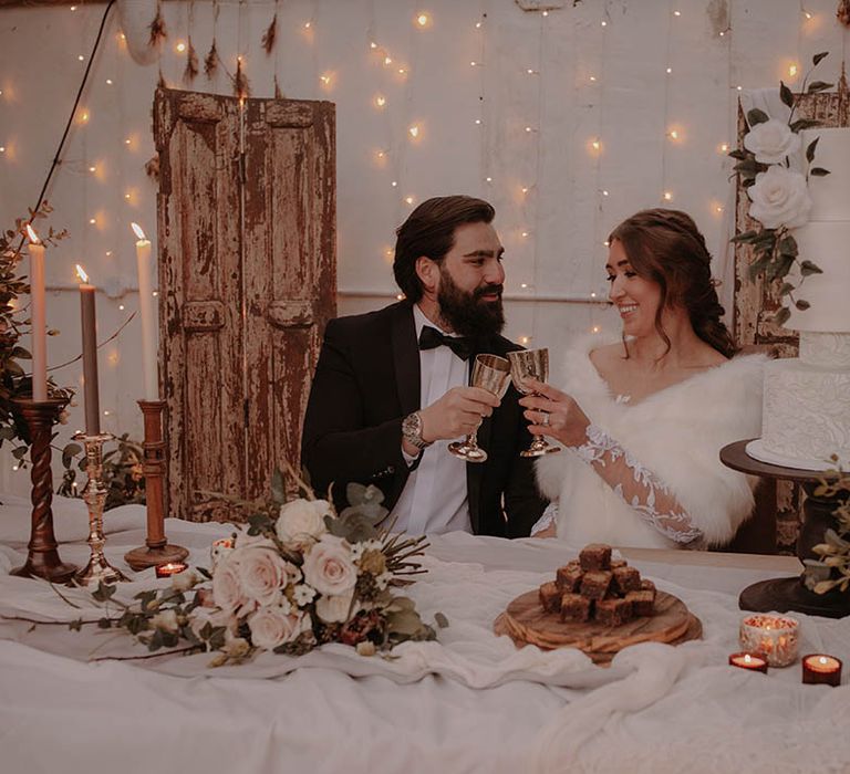 Bride and groom cheers their gold goblets next to their textured white wedding cake for their rustic winter wedding 