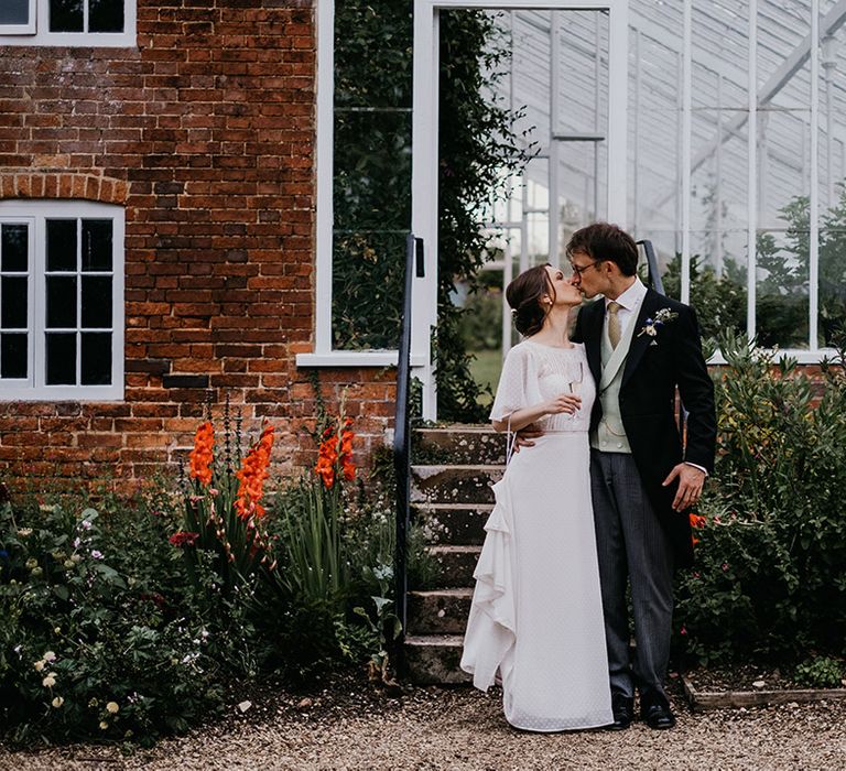 Bride in batwing sleeve lace detail wedding dress and groom in three piece suit with light waistcoat holding champagne glasses kissing outside Kelmarsh Hall