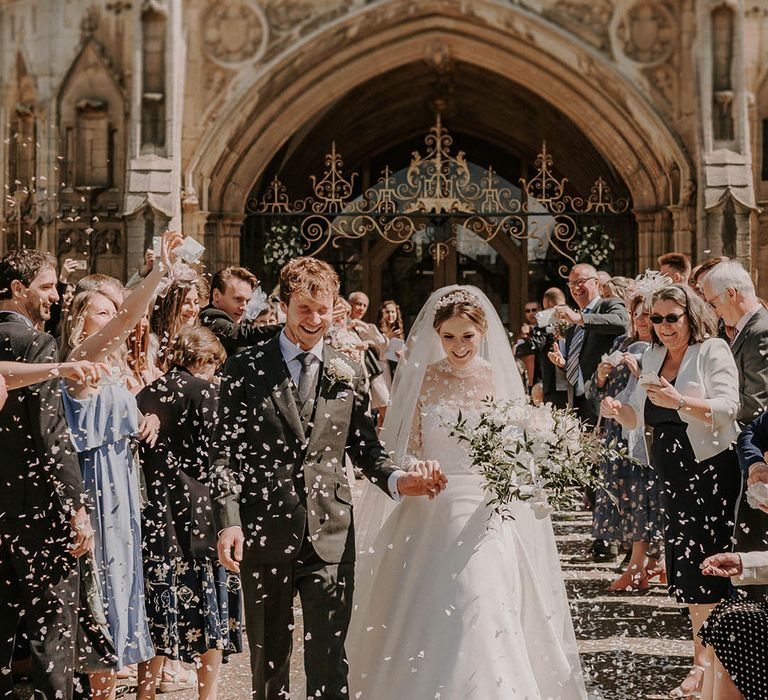 Groom in a black three piece suit walks holding hands with the bride as they have a confetti exit 