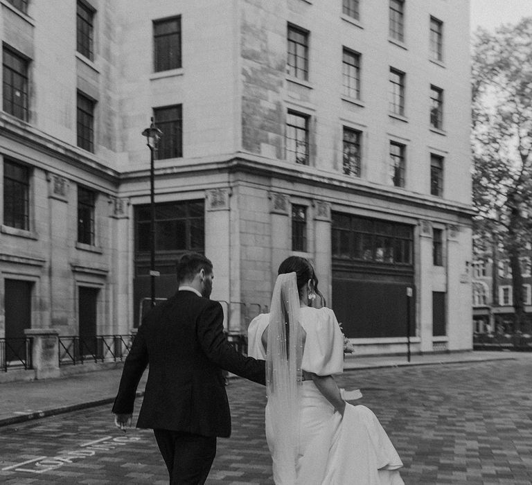 Bride in open back puff sleeve wedding dress with pearl detail veil and groom in dark tux walking hand in hand