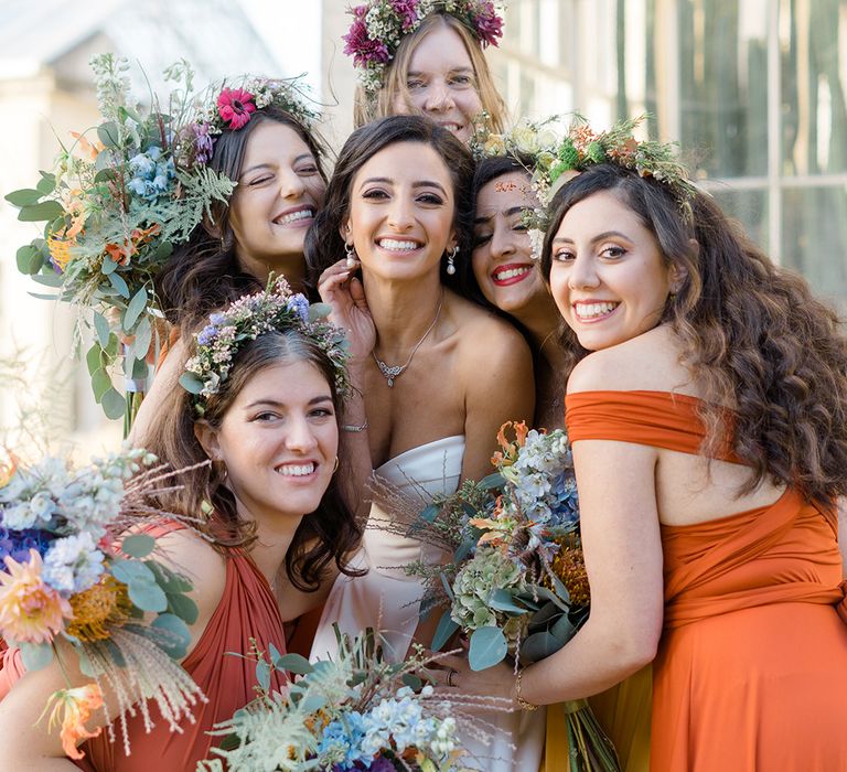 Bride stands with her bridesmaids who wear colourful ombre styled bridesmaid dresses in orange and yellow complete with floral crowns