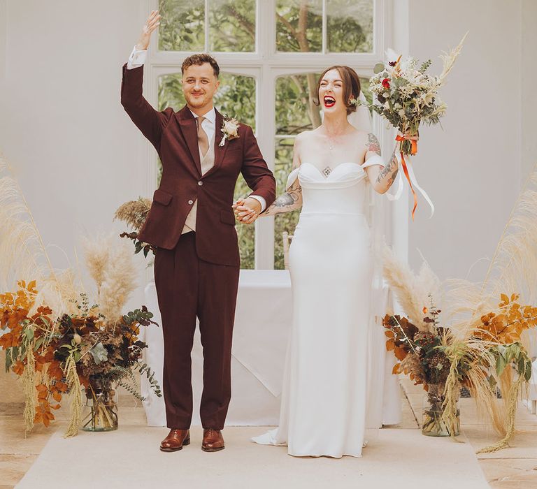 Bride holds up Autumnal styled bouquet and walks alongside her groom who wears maroon suit complete with floral buttonhole