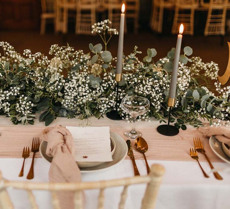 Neutral wedding tablescape with white table cloth, neutral pink table runner, sage green candles, minimal wedding menus, rose gold cutlery and foliage and baby's-breath table decorations