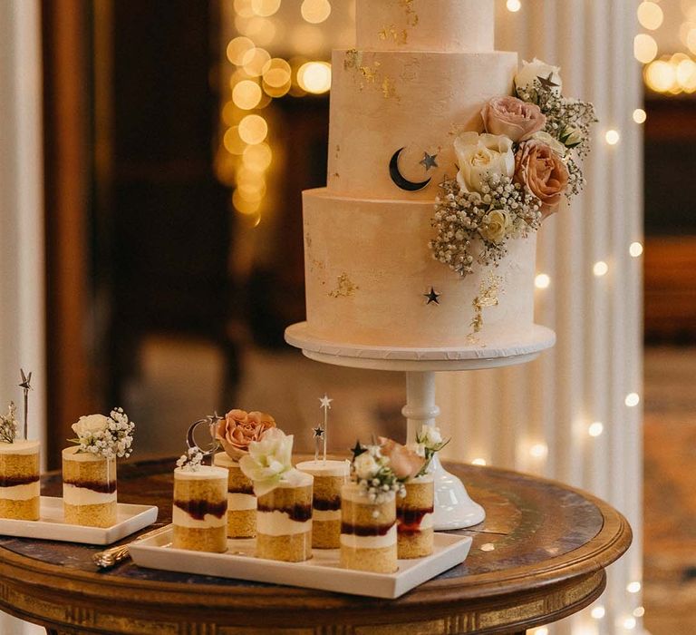 Three tiered buttercream wedding cake with neutral white and pink floral, foliage and star and moon decorations on white cake stand with mini Victoria sponge cakes