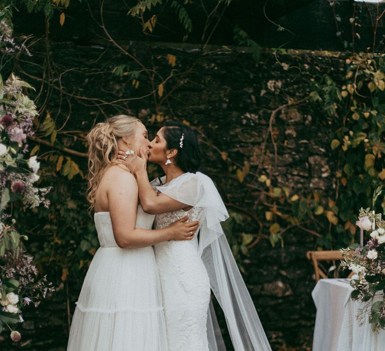 Brides in complimenting lace wedding dresses, one with short puff sleeves and the other strapless, kissing at the alter