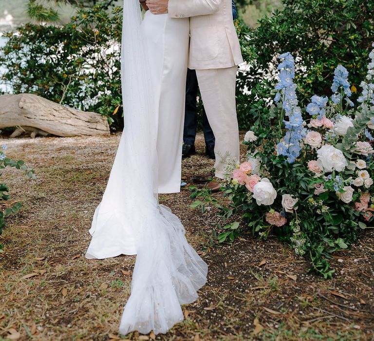 Bride and Groom kiss in front of trees whilst holding vows with Brides’ train and veil flowing along the ground