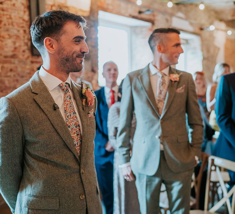 Groom smiles as he sees the bride for the first time in a tweed suit with a pink buttonhole and floral tie 