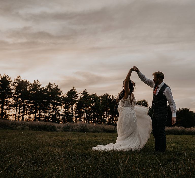 The groom in a grey checkered suit spins the bride as the couple spend some time outside during sunset for their couple portraits 