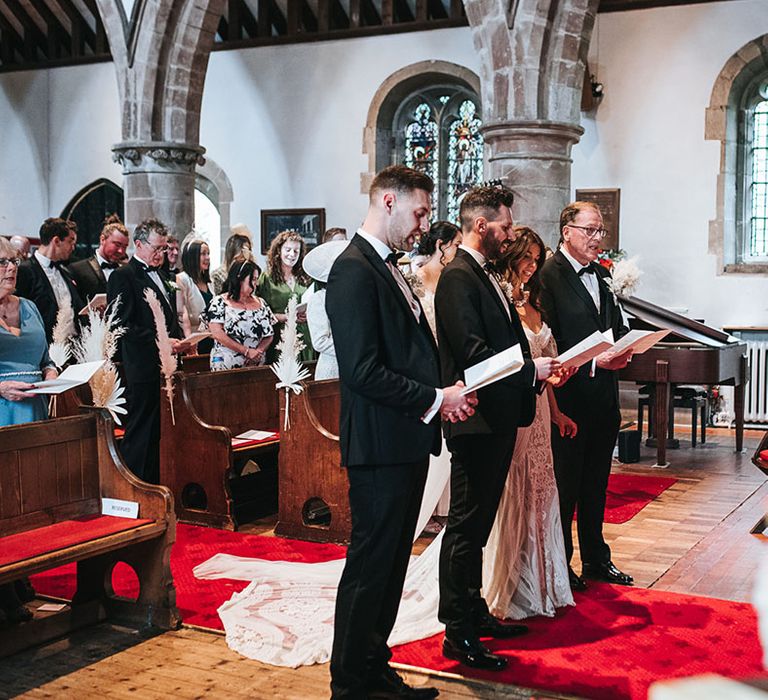 Groom, groomsmen, father of the bride and bride stand with order of service booklets listening to the officiant for the wedding ceremony 