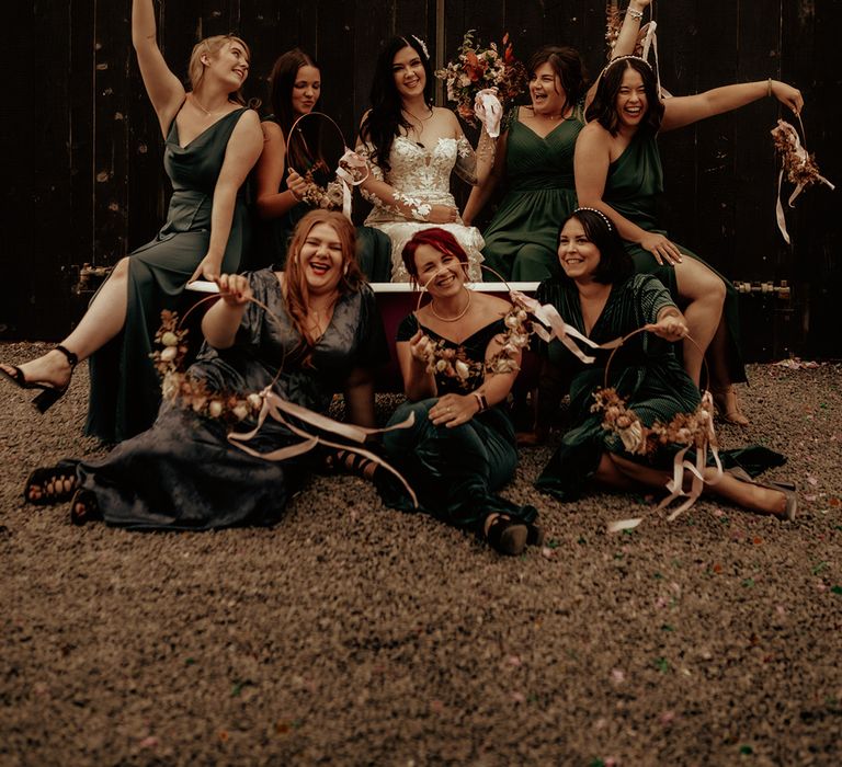 Bridal party posing in copper bathtub at The Giraffe Shed wedding venue wearing mismatched bridesmaid dresses and bride wearing off the shoulder bodycon wedding dress with long, sheer sleeves and lace detailing holding dried floral bouquet and dried floral wreaths