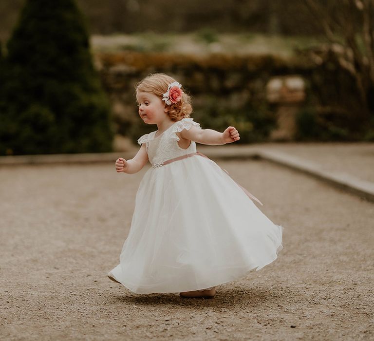 Flower girl wears white floor-length dress with flowers in her hair and pale pink embellished belt