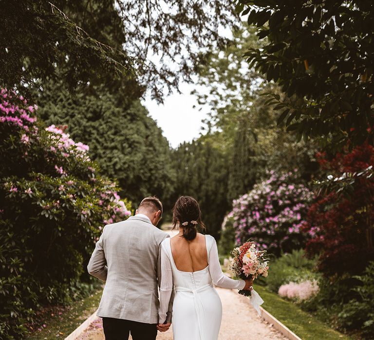 Groom in black suit trousers and grey blazer holding hands with bride in square back detail long sleeve satin wedding dress with rose and foliage wedding bouquet