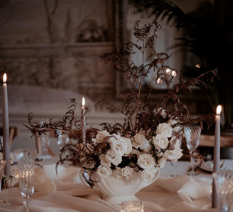 Minimalist wedding tablescape with beige tapered candles in clear candlestick holders and large textured white ceramic vase with white rose and dark dried flower arrangements 