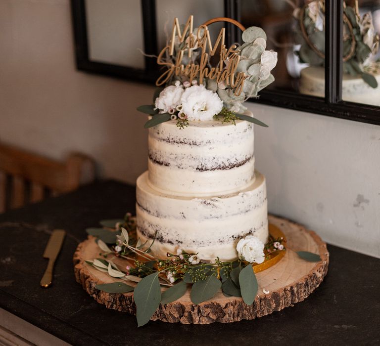 White frosted naked wedding cake complete with floral decor and gold personalised cake topper