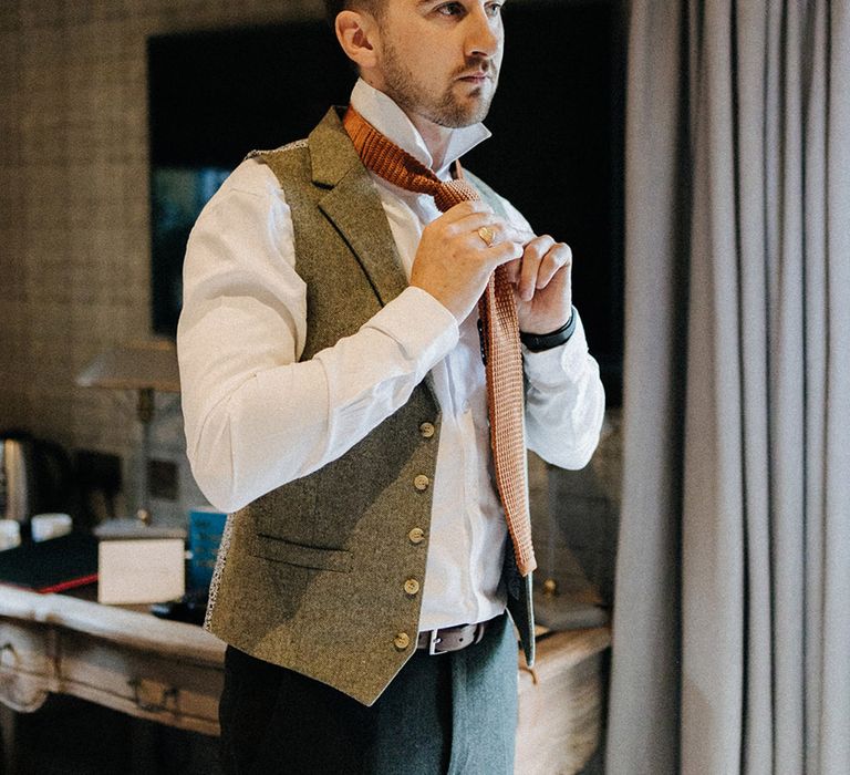 Groom in a white shirt and grey tweed waistcoat does up his orange tie for the wedding day 