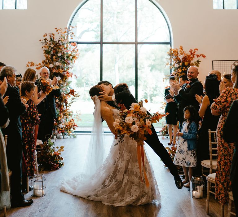 Groom dips his bride for kiss as she holds burnt orange bridal bouquet after bespoke wedding ceremony