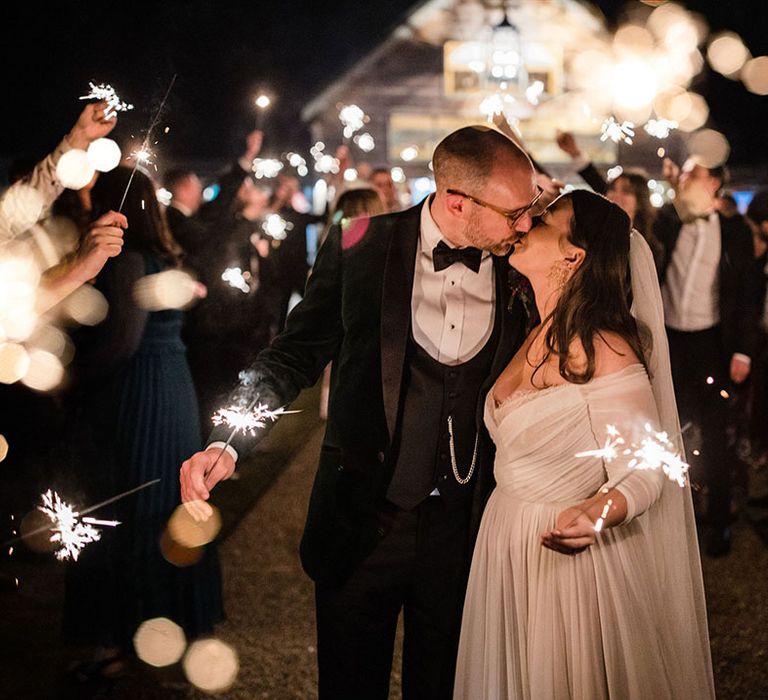 Groom in traditional black tie kissing the bride as they wave sparklers for their sparkler send off 