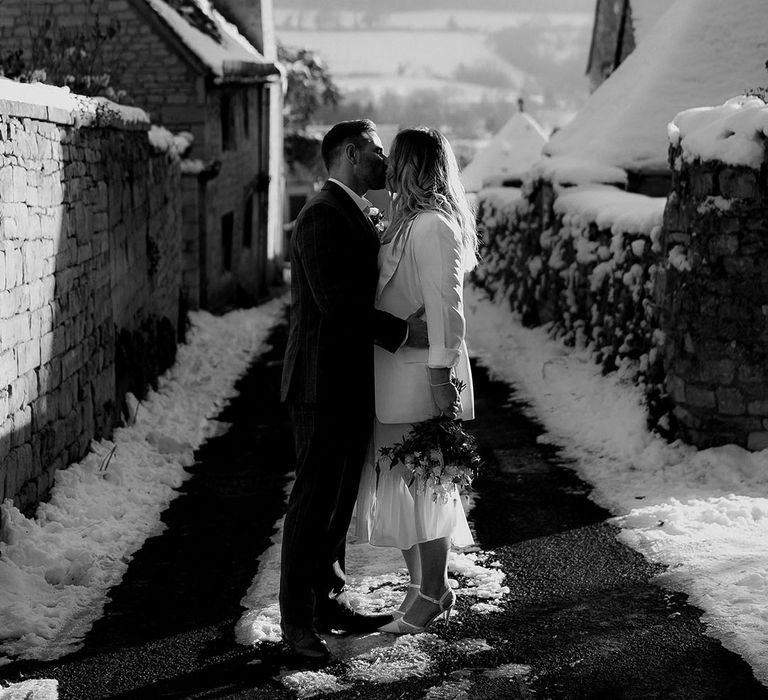 Groom in dark grey suit kissing the bride in white blazer cover-up for snowy Christmas wedding in the Cotswolds 