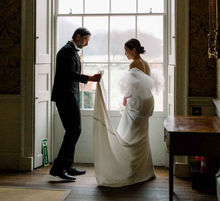 Bride in strapless wedding dress with puff tulle sleeves standing with groom in classic black tuxedo, black bowtie and boutonniere holding her train