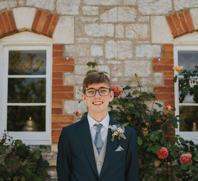 The groom in a navy suit jacket with light grey waistcoat with light blue tie and daisy buttonhole with glasses 