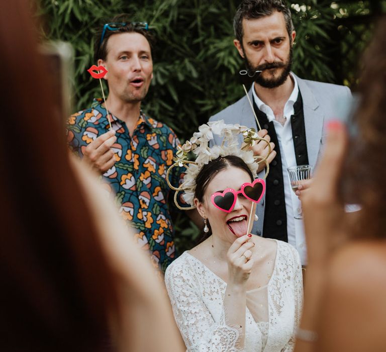 Bride in white bridal crown holding loveheart sunglasses prop with guests at wedding photobooth