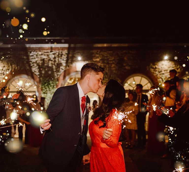 Bride in red lace wedding dress with long sleeves, red and gold hair accessories and golden closed toe wedding heels doing wedding sparkler exit with groom in deep blue grooms blazer, grey waistcoat, red tie, red pocket square and mixed dried flower boutonniere at The Orangery Maidstone