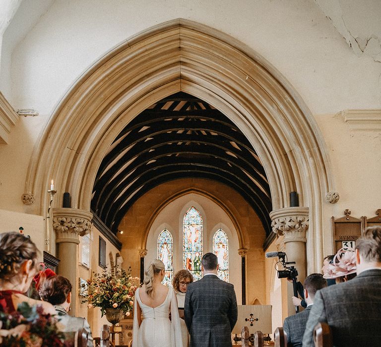 Bride in Karen Willis Holmes wedding dress with tulle wings standing at the altar with the groom in a three piece grey suit for their church ceremony 