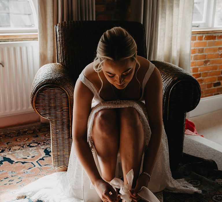 Bride sitting down putting on her white wedding shoes tied up with white ribbon straps