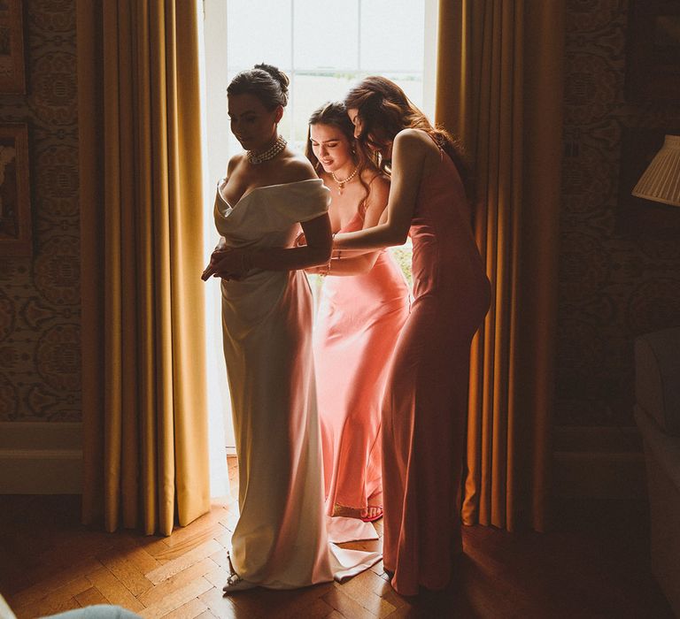 Bridesmaids in satin pink bridesmaid dresses stand with the bride helping with the final touches for the day 