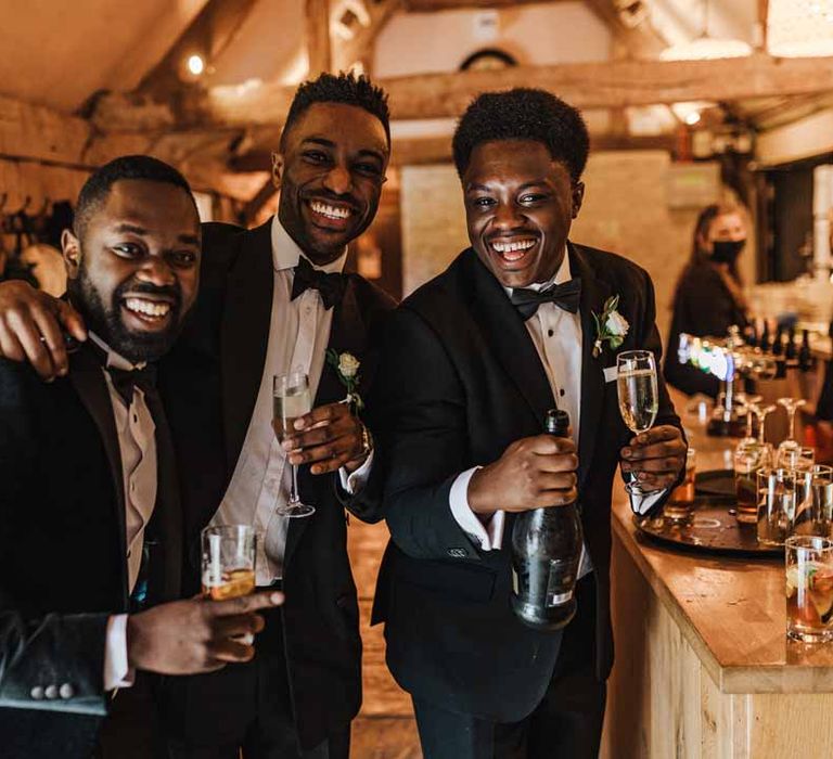 Groomsmen wearing classic black suits with bowties and white garden rose and foliage boutonnieres holding glasses of champagne and smiling at Irish-Ghanaian fusion wedding