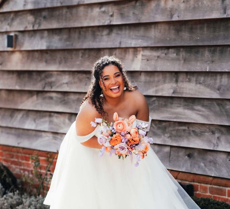 Bride in off the shoulder wedding dress holding orange, lilac and peach mixed flower bridal bouquet and laughing at Rackleys Barn