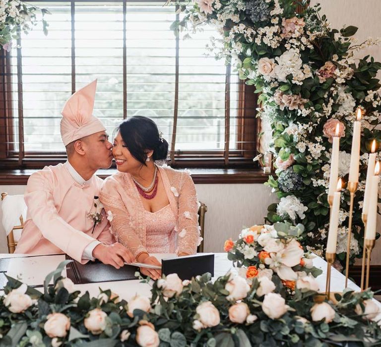 Bride in floral 3D applique pink and rose gold htamane sitting with groom in baby pink htaingmathein signing the register at Burmese wedding underneath floral wedding bouquet 