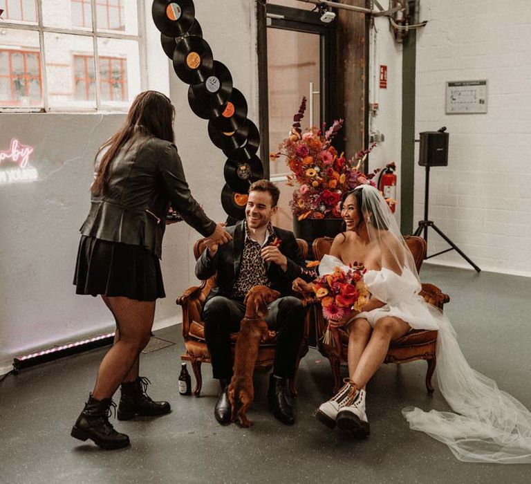 Wedding celebrant wearing leather jacket greeting bride and groom sat by vinyl wedding arch in crushed velvet armchairs 