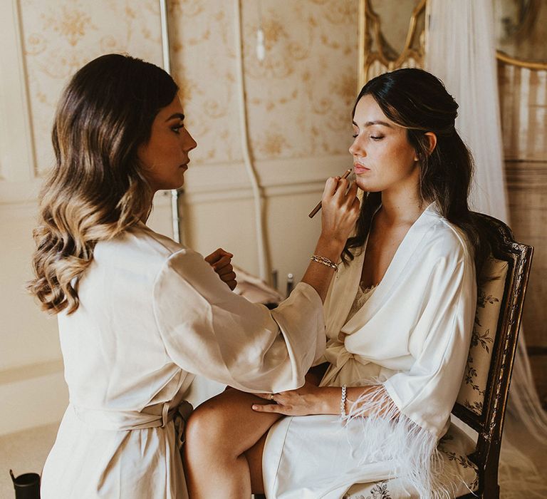 Bride in white satin wedding robe sitting on a chair getting her makeup done for the wedding day