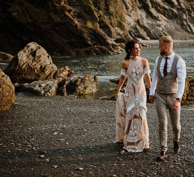 Bride in Rue de Seine wedding dress walking along the beach with the groom in a grey suit