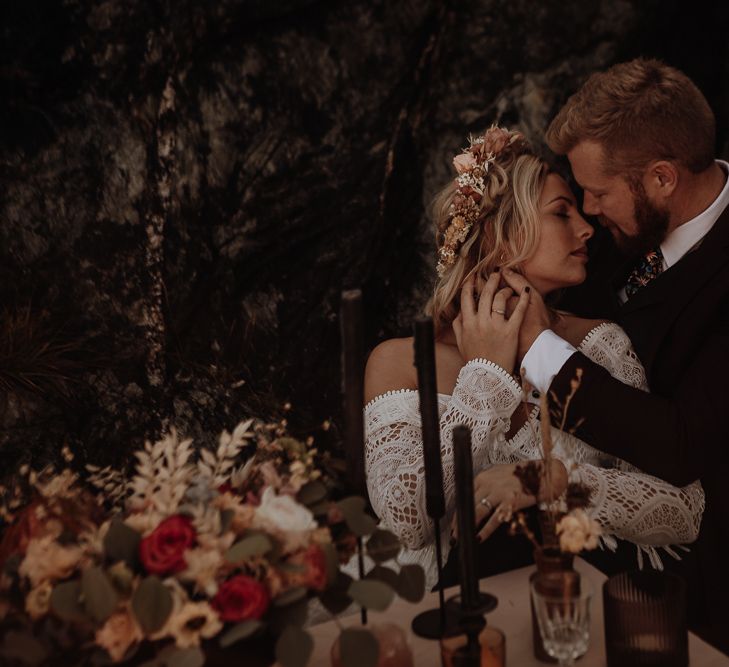 Bride in boho lace wedding dress posing with groom next to private table setting with black candles and a flower crown 