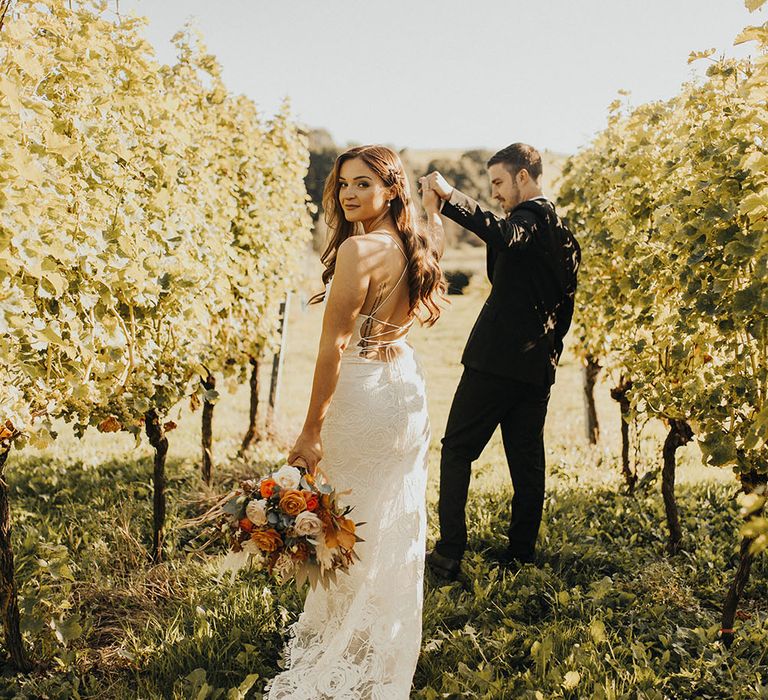 Bride wears backless cowl neck wedding dress as she walks through vineyard with groom