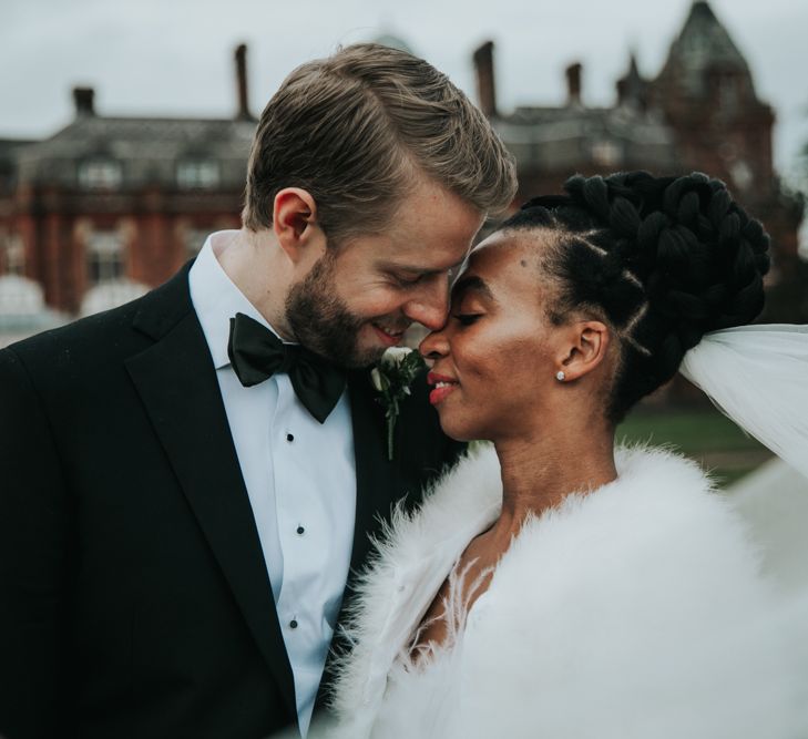 A bride and groom snuggle into each other. tHe bride wears her afro hair in twists that are positioned in an updo on top of her head. She wears a fluffy cover up over her shoulders.