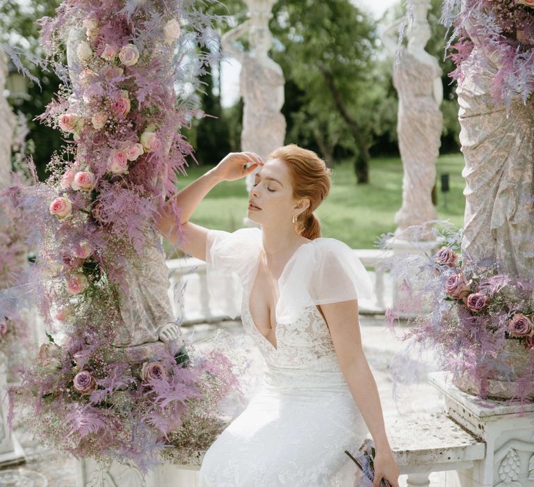 Ginger haired bride sits surrounded by lilac floral bouquets 