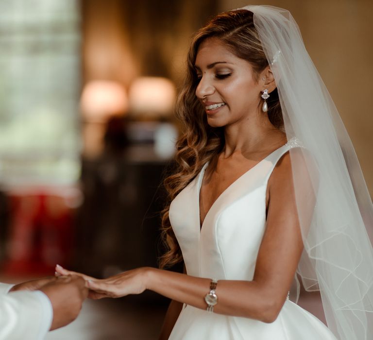 Bride in a fitted bodice wedding dress with side swept wavy hair and cathedral length veil putting on her wedding ring during the ceremony 