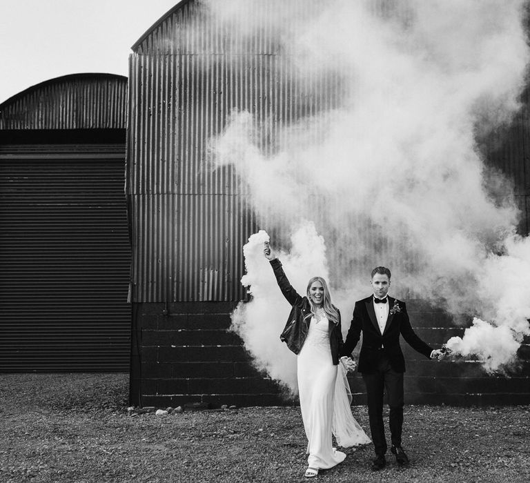 Bride in personalised leather bridal jacket and white Charlie Brear wedding dress holding hands with groom in black Hugo Boss suit holding white smoke bombs