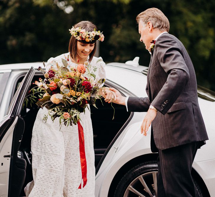 Bride getting our of the white wedding car in a white Broderie Anglaise wedding dress with colourful flower crown and bouquet 