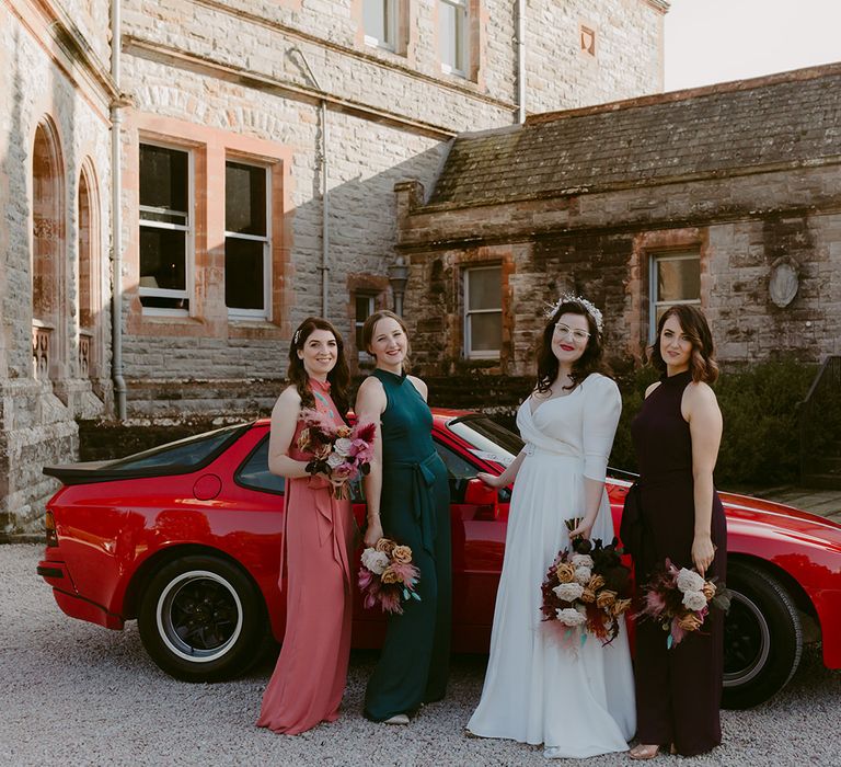 Bride in Rime Arodaky dress with bridesmaids in different coloured Rewritten bridesmaids dresses holding bouquets in front of red vintage sports car at Castle Leslie wedding