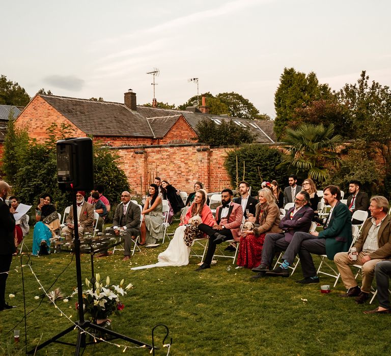 Wedding party gather outdoors and sit listening to speeches